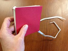 a hand holding a pink book next to a white piece of paper on top of a wooden table