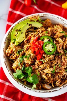 a white bowl filled with shredded meat and topped with avocado, red peppers, jalapenos, and cilantro