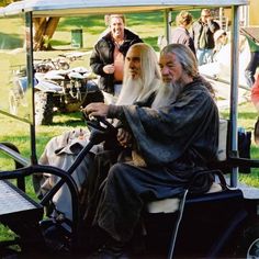 an old man and woman sitting on a golf cart in the grass with other people behind them