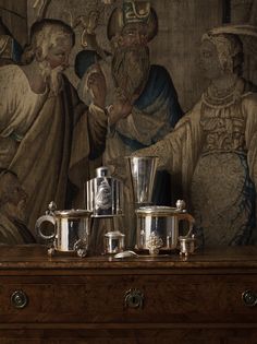 silver coffee pots and cups sit on a table in front of a painting with figures