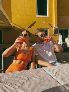 a man and woman sitting next to each other drinking drinks from glasses in front of a yellow building