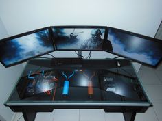 three computer monitors sitting on top of a glass table with video game controllers in front of them