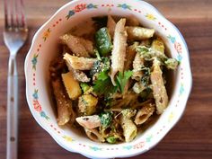 a white bowl filled with pasta and vegetables next to a fork on top of a wooden table