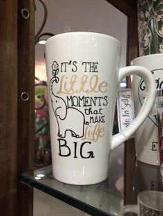 two coffee mugs sitting on top of a glass shelf next to each other, with the words it's the little moments that make life big