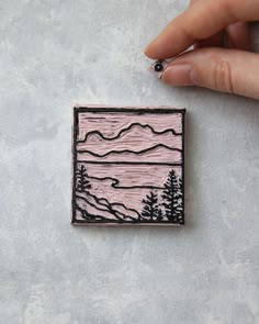 a hand holding a small wooden stamp with mountains in the distance and trees on it