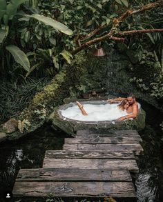 a woman laying in a hot tub surrounded by greenery and trees on a wooden platform