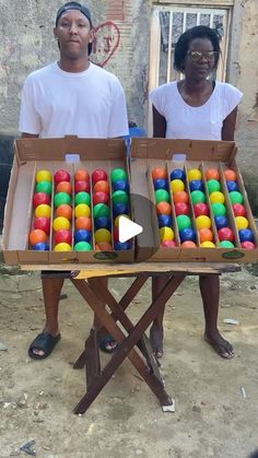 two people standing next to each other in front of an open box filled with colorful balls