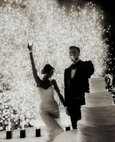 a bride and groom are standing next to a wedding cake with fireworks in the background