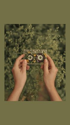 two hands holding up an old fashioned cassette in front of some plants and trees, with one person's hand on top of the cassette