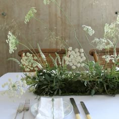 the table is set with white flowers and greenery on it, along with silverware