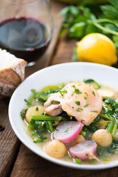 a white bowl filled with food next to a glass of wine and bread on top of a wooden table