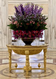 a large vase filled with lots of purple flowers on top of a gold table in a room