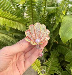 a hand holding a colorful flower brooch in front of green plants and leaves on the ground