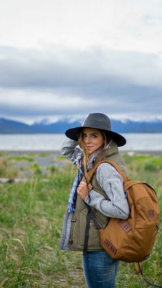 a woman wearing a hat and carrying a brown backpack in the grass with mountains in the background