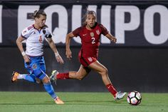 two women playing soccer on a field with the ball in front of them and one running after it