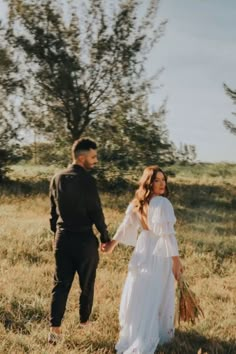 a man and woman holding hands while walking through a field with trees in the background