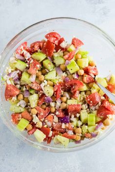 a glass bowl filled with chopped vegetables and chickpeas, on top of a white surface