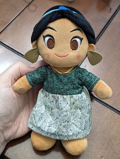 a hand holding a small stuffed doll with brown eyes and dark hair, wearing a green dress