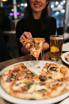 a woman eating a slice of pizza at a table with a glass of beer in front of her