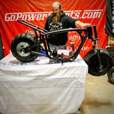 a man standing next to a motorcycle on top of a white cloth covered table in front of a red banner