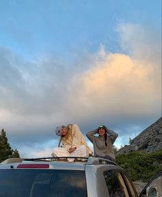 two people sitting on the roof of a car in front of a mountain with clouds