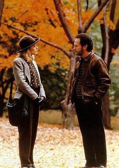 two people standing next to each other in front of trees with leaves on the ground