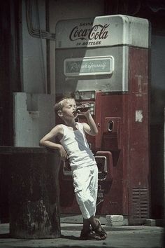 a young boy standing next to an old coca cola machine and talking on the phone