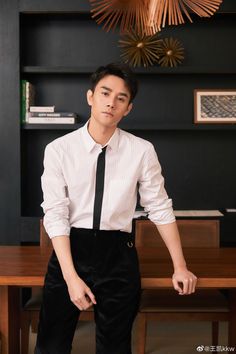 a young man wearing a white shirt and black tie sitting at a wooden table in front of a bookcase