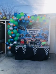 a table topped with balloons next to a monster truck birthday party sign and checkered table cloth