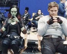 two people dressed as aliens sitting in chairs with cell phones and looking at the camera