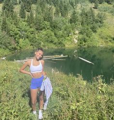 a woman standing in front of a lake with trees on the hillside behind her and mountains in the background