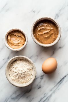 three bowls filled with different types of food next to an egg and flour on a marble countertop