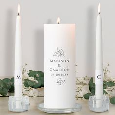 two white candles sitting next to each other on top of a wooden table with greenery