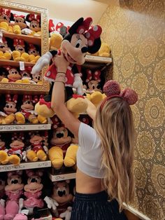 a woman holding onto a mickey mouse stuffed animal in front of a wall full of other stuffed animals