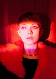 a woman with short hair wearing a red fur collar and black top, standing in front of a wooden wall