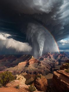 a rainbow is seen in the sky above a large, dark cloud over a canyon