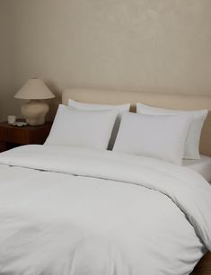 a bed with white sheets and pillows in a bedroom next to a lamp on a night stand