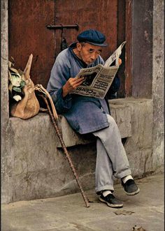 an old man sitting on a ledge reading a newspaper