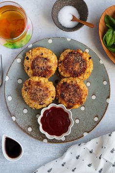 three crab cakes on a plate with ketchup, sauce and spoons next to it