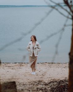 a woman standing on top of a sandy beach next to the ocean wearing a coat