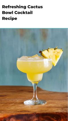 a glass filled with a yellow drink sitting on top of a wooden table next to a slice of pineapple