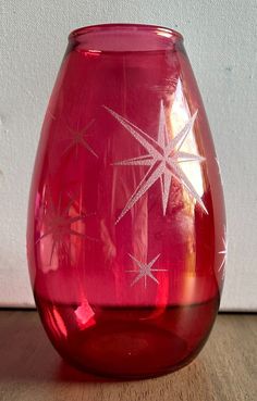 a red glass vase sitting on top of a wooden table next to a white wall