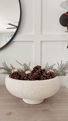 a white bowl filled with pine cones sitting on top of a wooden table next to a mirror