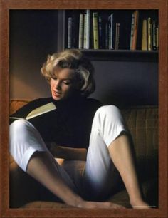 a woman sitting on a couch reading a book with her legs crossed and wearing white pants