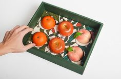 a person reaching for some fruit in a green box on a white table with oranges and apples