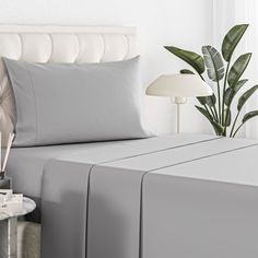 a bed with grey sheets and pillows next to a plant in a vase on a table