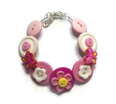 "Materials: New and recycled buttons, craftwire, metal lobster claw clasp. Length:  The bracelet measures 18cm (7\") but can be made longer if the chain is fully extended.  (maximum length 9\" or 22cm) This handmade bracelet is made of new and re-purposed buttons.  It features two pink butterflies sitting amongst a field of flowers and pink buttons.  It has been strung on a double strand of tarnish-resistant silver craft wire, which can be moulded to suit the wearer's wrist. There is a lobster claw clasp and the largest button is approximately 1\"  (2.5 cm). This pretty, affordable piece of jewelry is sure to bing lots of comments and admiring glances. All my button jewellery is unique and hand made by me. It would make a very special gift for anyone who is over the age of 5, as small butt Butterflies Bracelet, Button Necklaces, Recycled Buttons, Bracelet Butterfly, A Field Of Flowers, Wrist Accessories, Button Necklace, Button Bracelet, Field Of Flowers