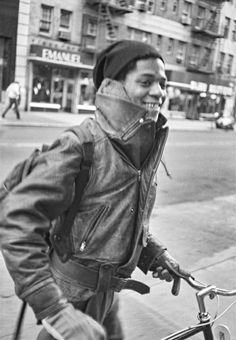 a black and white photo of a man riding a bike down the street with a hat on