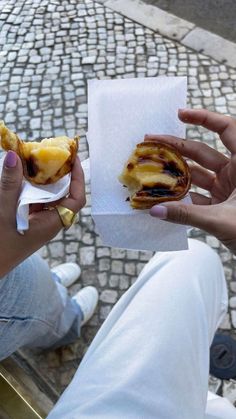 two people holding food in their hands while sitting on a bench outside with cobblestone pavement