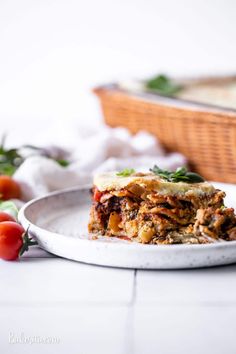 a white plate topped with lasagna covered in sauce and toppings next to a basket of tomatoes